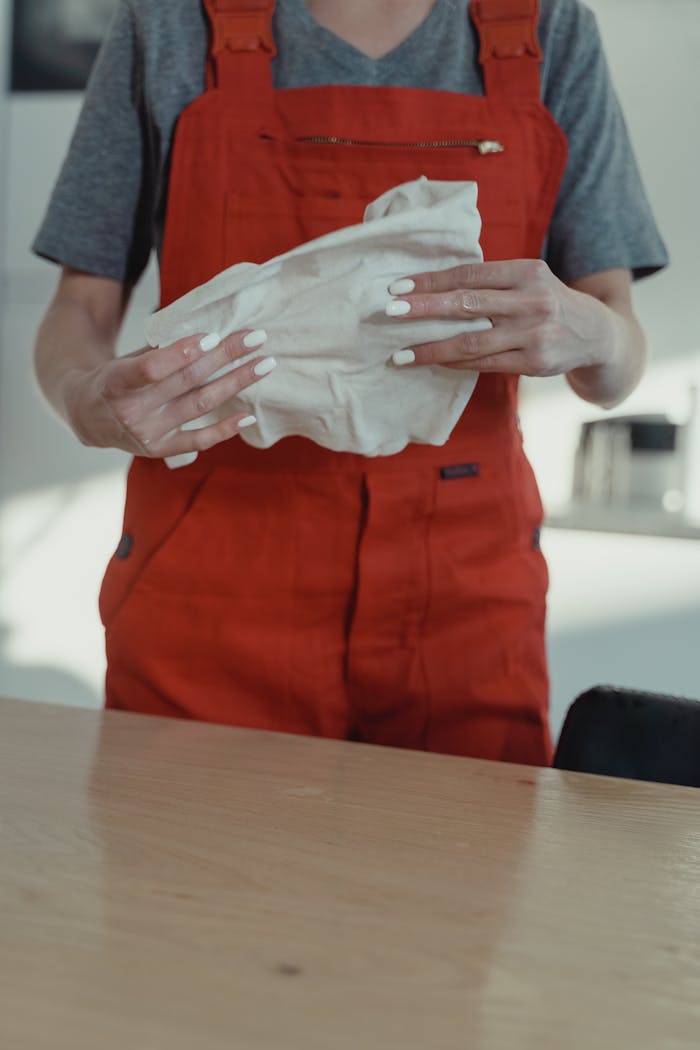 A woman wearing red overalls cleans a wooden table with a white cloth indoors.